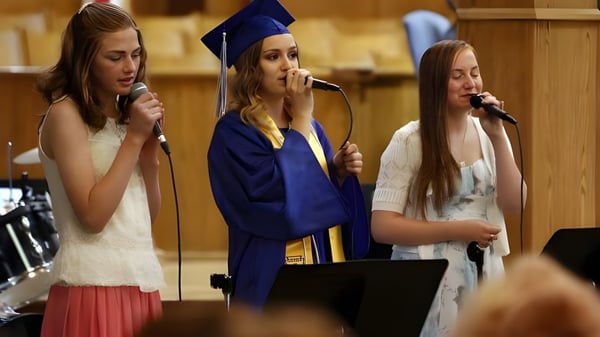 Tres estudiantes de la Immanuel Christian Secondary School cantan en togas de graduación en un escenario con micrófonos.
