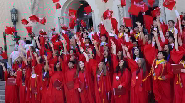 Estudiantes de la Incarnate Word Academy se reúnen en togas de graduación rojas en las escaleras de un edificio.