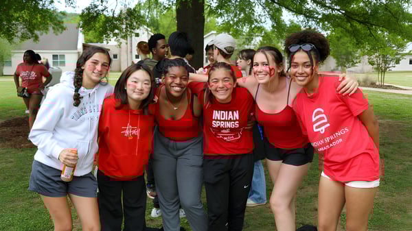 Un grupo de jóvenes en camisetas rojas de la Indian Springs School está afuera en un campo con árboles de fondo.