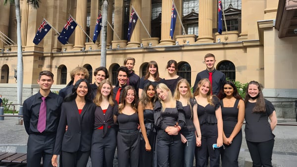 Un grupo de estudiantes de la Indooroopilly State High School está de pie en ropa formal frente a un gran edificio con columnas y banderas australianas.