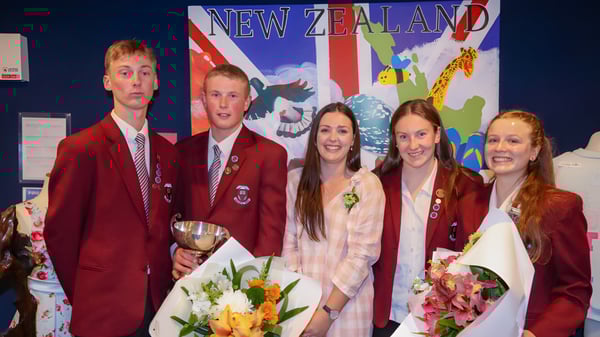 Un grupo de estudiantes de Inglewood High School en uniformes rojos está con flores y un trofeo frente a una gran bandera neozelandesa.