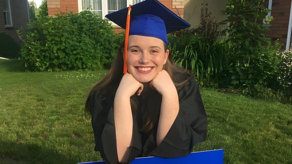 Una graduada sonriente en toga está de pie en el campus de la Innisdale Secondary School frente a un edificio de ladrillos al aire libre.