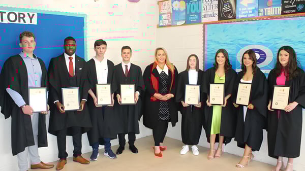 Estudiantes del Inver College en togas de graduación sostienen sus certificados frente a una pared colorida con carteles.