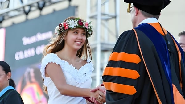 Una estudiante de la Iolani School lleva un vestido blanco y una corona de flores y le da la mano a un hombre en toga académica frente a un edificio universitario.