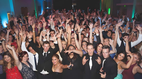 Un gran grupo de estudiantes de la Ipswich State High School levantan las manos en una sala de eventos iluminada de colores.