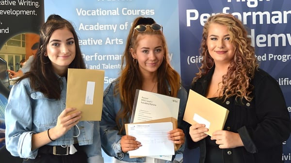 Tres alumnas sostienen sus certificados frente a un fondo informativo sobre el Itchen Sixth Form College.