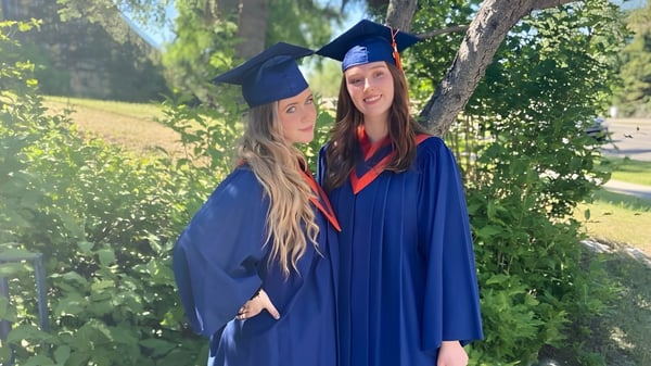 Dos estudiantes de la James Fowler High School están al aire libre en sus vestidos de graduación con árboles de fondo.