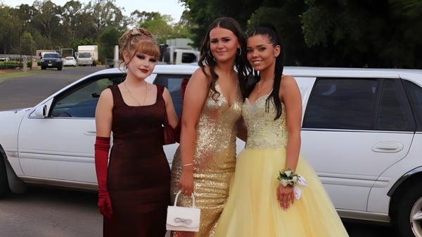 Tres estudiantes de la James Nash State High School están vestidas de forma formal frente a un vehículo blanco en el área exterior de la escuela.