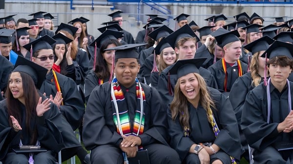 Un gran grupo de graduados en togas y birretes negros durante la ceremonia de graduación de Jeffco Public Schools al aire libre.
