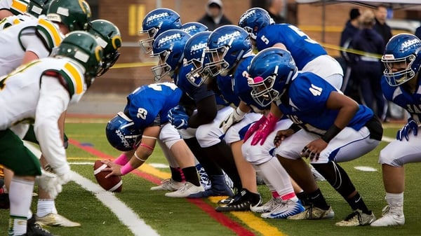 Estudiantes del John Abbott College están en camisetas azules y verdes en el campo y se preparan para una jugada de fútbol americano.
