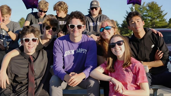 Un grupo de estudiantes de la John Bapst Memorial High School posa afuera frente a un colorido fondo de árboles y cielo.