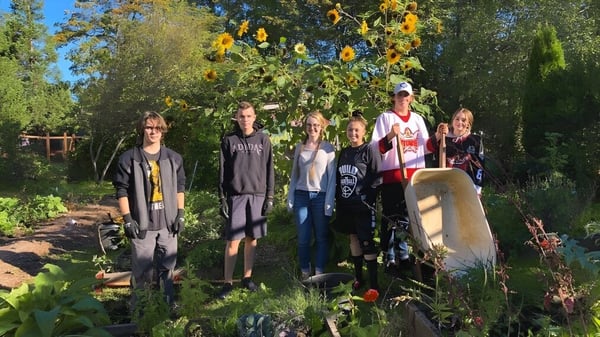 Un grupo de personas está de pie en un jardín colorido con árboles y una escultura de piedra en el terreno de la John Barsby Secondary School.