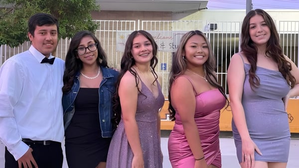 Un grupo de alumnas y alumnos se encuentra frente a un edificio con una valla de metal en el terreno de la John F. Kennedy High School.