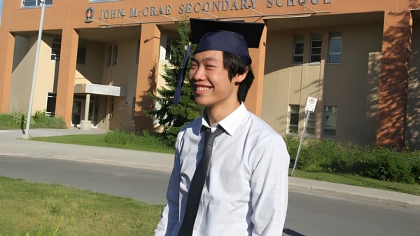 Un estudiante de la John McCrae Secondary School está en toga y birrete frente al edificio escolar.