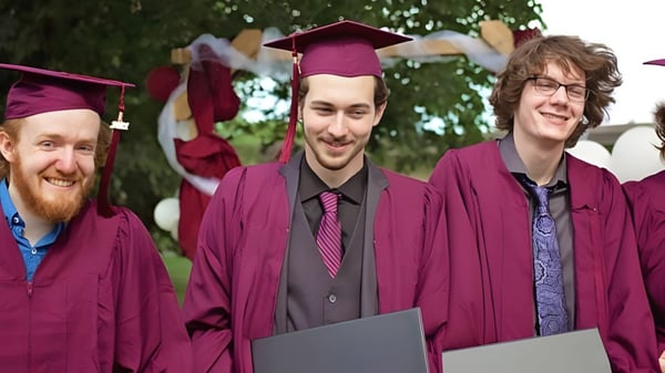 Tres hombres en ropa de graduación están juntos al aire libre en el terreno de la John McGregor Secondary School.