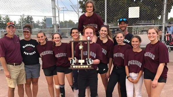 Estudiantes de la John McGregor Secondary School celebran la victoria de un trofeo frente a un campo deportivo.