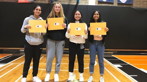 Cuatro estudiantes de la John Oliver Secondary School están en el gimnasio y sostienen carteles amarillos frente a sus rostros.