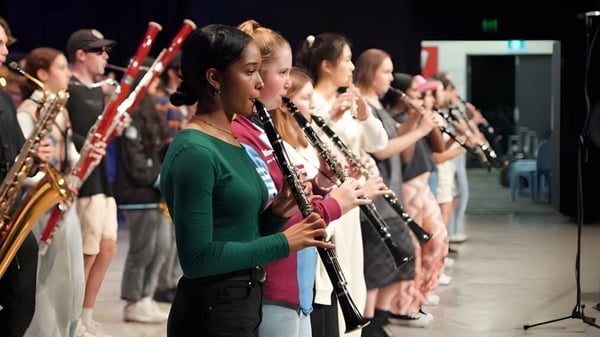 Estudiantes del John Paul College tocan instrumentos de viento en un escenario dentro de una orquesta.