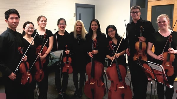 Un grupo de músicos con violines y cellos se encuentra juntos en una sala del John Polanyi Collegiate Institute.
