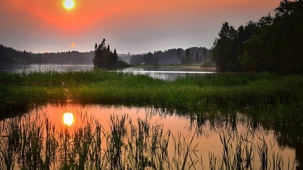 Un lago tranquilo con densos bosques verdes y un cielo anaranjado se refleja en el terreno de la John Rennie High School.