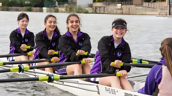 Un grupo de alumnas de la John Scottus Secondary School está sentada en un bote de remo sobre el agua con vista a la ciudad de fondo.
