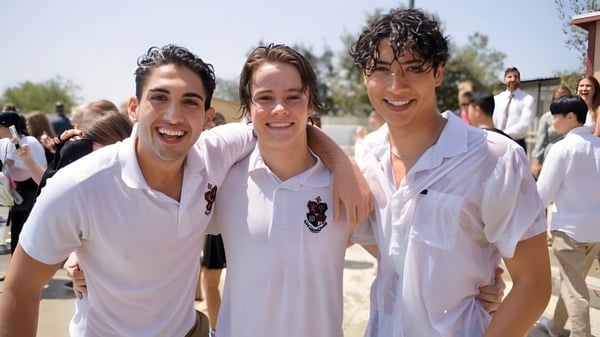 Tres estudiantes de la JSerra Catholic High School están sonriendo al aire libre frente a árboles y otras personas.