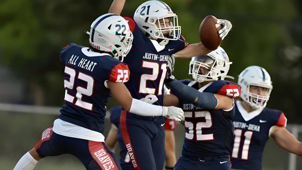 Un grupo de jugadores de fútbol americano de la Justin Siena High School juega en un campo de césped bajo un cielo azul.