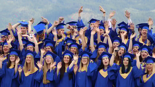 Los graduados de la Kalamalka Secondary School celebran en togas azul-amarillas su graduación frente a un paisaje montañoso.