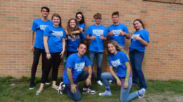 Un grupo de estudiantes de la Kalkaska High School está de pie frente a una pared de ladrillo y lleva camisetas azules uniformes.