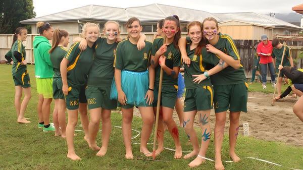 Un grupo de jóvenes atletas de Katikati College está en un campo de hierba frente a un edificio residencial.