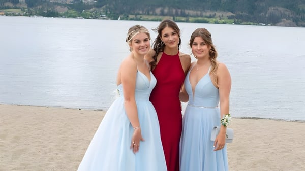 Tres estudiantes de la Kelowna Secondary School están vestidas de manera formal en la playa con un lago y montañas de fondo.
