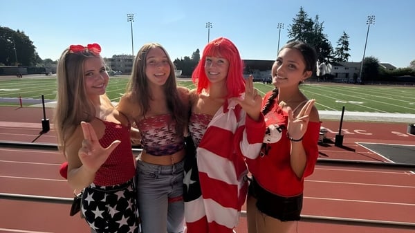 Cuatro estudiantes de la Kennedy Catholic High School posan con ropa patriótica en un campo deportivo con un estadio al fondo.