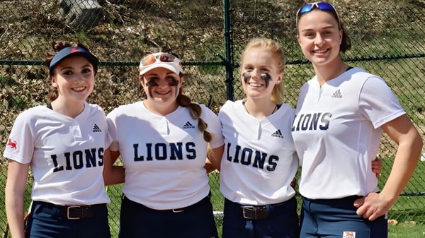 Cuatro estudiantes de la Kent School están de pie en camisetas blancas de leonas frente a un campo deportivo cercado.