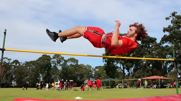 Una alumna del Kiara College salta al aire libre sobre una barra de salto de altura frente a árboles y otras personas.