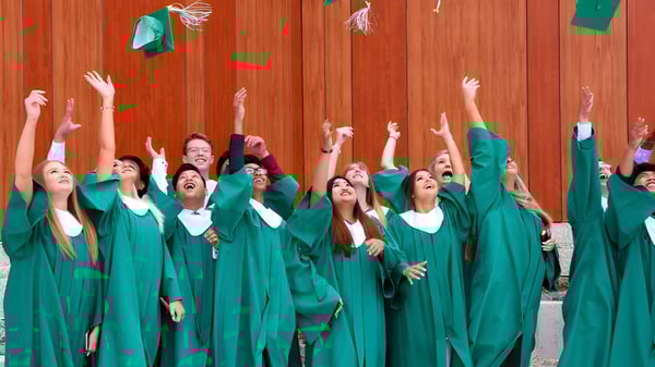 Los graduados de Kildonan East Collegiate en togas verdes celebran frente a una pared de madera.