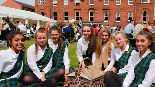 Un grupo de alumnas de la Kilgraston School lleva ropa tradicional escocesa frente a un área de carpas con más personas.
