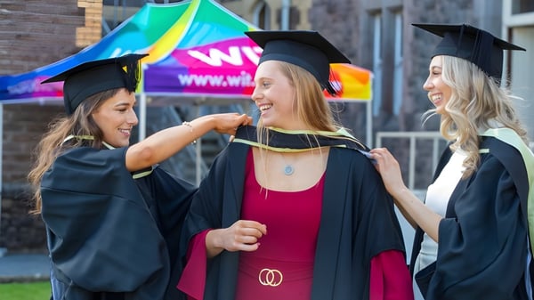 Tres alumnas de la Kilkenny City Vocational School celebran su graduación frente a un fondo colorido con un cartel de WIN.