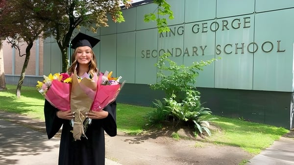 Una graduada está frente al edificio verde de la King George Secondary School sosteniendo un ramo de flores.