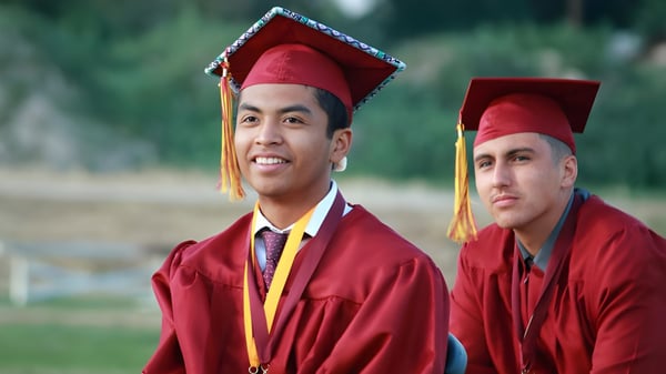 Dos egresados de la Kings Christian School llevan togas y sombreros rojos y están al aire libre en un prado con árboles de fondo.