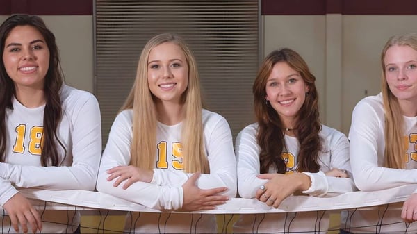 Cuatro estudiantes de la Kings Christian School en camisetas blancas con el número 13 están juntas en un entorno deportivo frente a una cerca de metal.