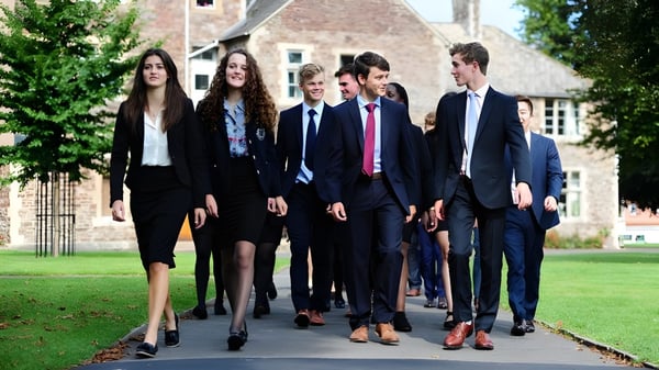 Un grupo de estudiantes del King's College camina por un camino entre arbustos verdes cerca de un edificio histórico.