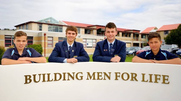 Cuatro estudiantes de la King's High School están sentados frente al cartel 'BUILDING MEN FOR LIFE' con el edificio escolar al fondo.