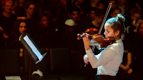 Una estudiante de la King's School Rochester toca el violín en el escenario frente a un público débilmente iluminado.