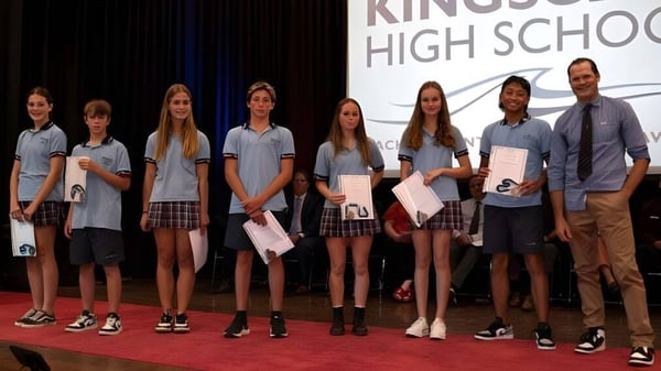 Un grupo de estudiantes está en el escenario de la Kingscliff High School sosteniendo certificados o premios.