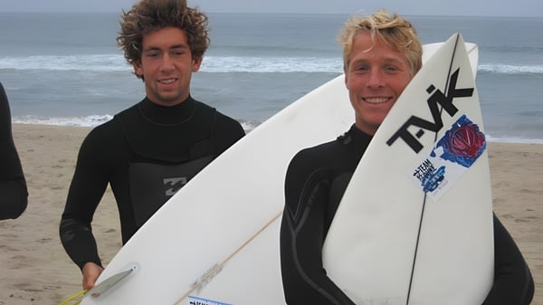 Dos estudiantes de la Kingston High School están de pie con tablas de surf en la playa frente al mar y un cielo nublado.
