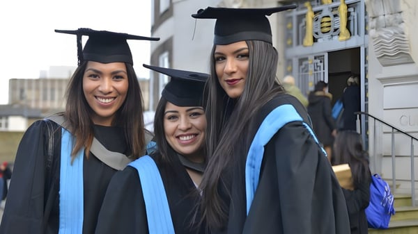 Tres estudiantes de la Kingston Secondary School en togas sonríen y posan frente a un edificio durante la ceremonia de graduación.