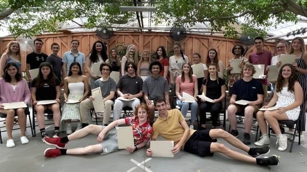 Un grupo de estudiantes de la Kingsville District High School se reúne al aire libre frente a una estructura de madera y vegetación verde.