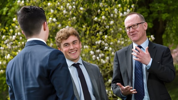 Tres hombres en trajes mantienen una conversación frente a un fondo verde en el campus de la Kingswood School.