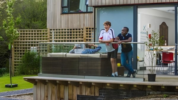 Dos personas están de pie en un deck de madera al aire libre con vegetación verde en el campus de la Kingswood School.