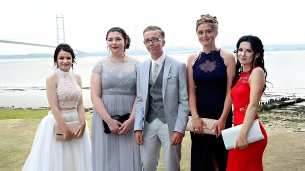 Un grupo de personas vestidas elegantemente posan al aire libre frente a un paisaje de agua y naturaleza en el terreno de la Kingswood School.